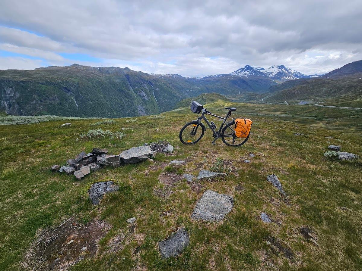 Bicycle touring with orange panniers on grassy mountain overlook with snow-capped peaks.