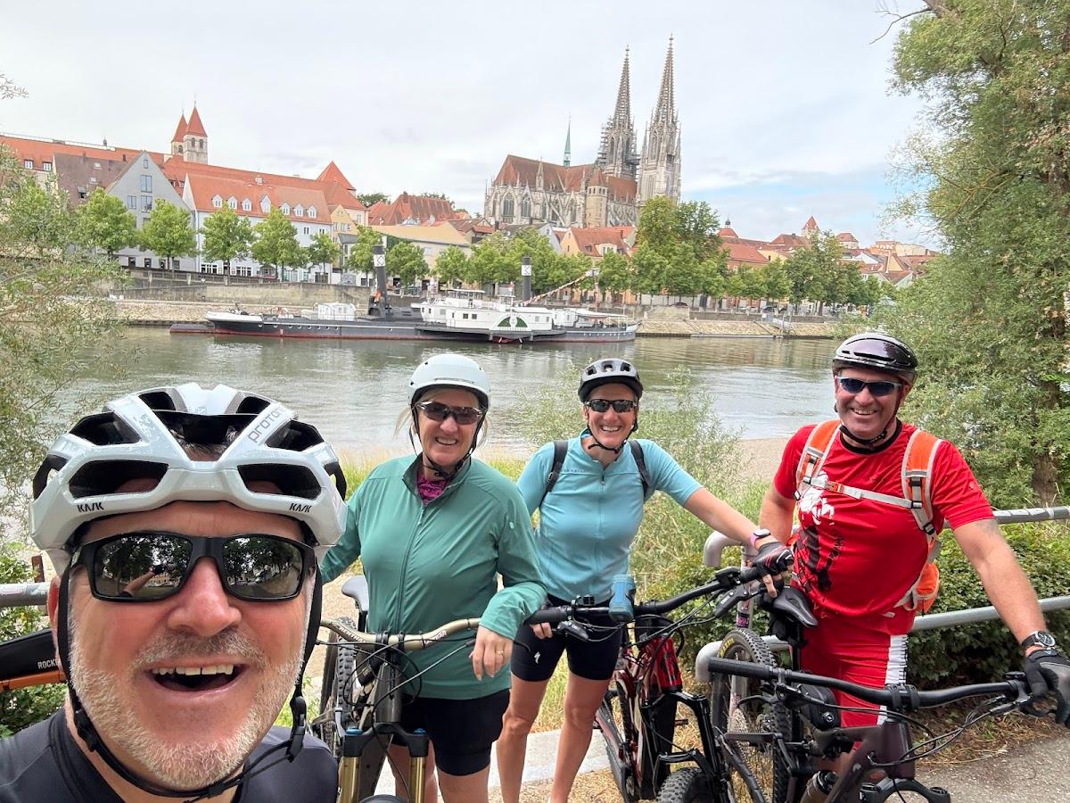 Cyclists taking a selfie by a river with a historic European city skyline and cathedral in the background.