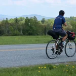 Mann auf dem Fahrrad auf einer Landstraße mit Bergen im Hintergrund (ohne Gesicht, wegschauend, Schotterfahrrad mit Bikepacking-Taschen) Reisen, Touren, Radfahren, Radfahren, Fahrrad